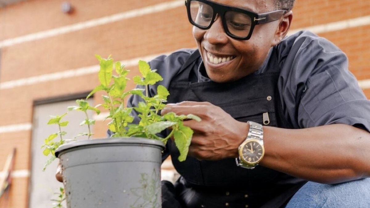 A chef crouching among potted herbs, holding a pot of basil
