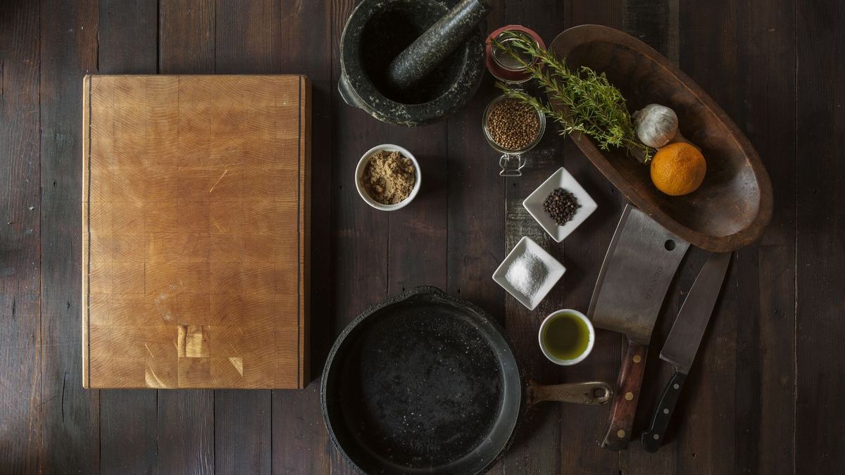 A chef's station from above: cutting board, cast-iron skillet, cleaver, mortar and pestle, herbs and spices on dark wood