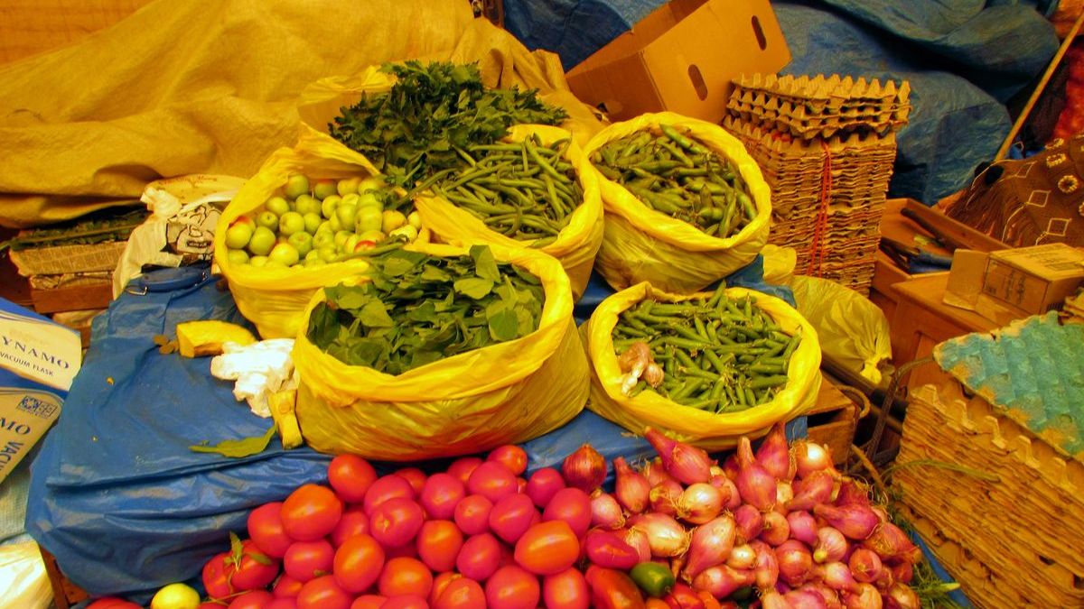 Sacks of leafy greens and beans over piles of tomatoes and shallots at a market stall